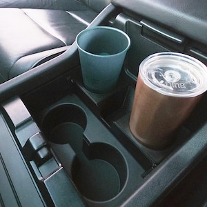 May include: A car's center console with two beverage holders and two cups. One cup is teal, and the other is brown with a clear lid. The console is black, and the car seat is black leather.