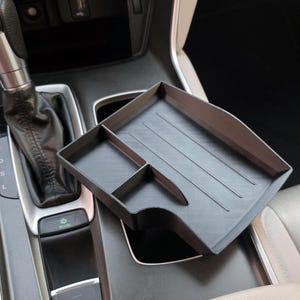 May include: A black car console organizer with multiple compartments. The organizer is designed to fit into a car's center console, providing storage for small items. The image shows the organizer in a car interior, near the gear shift.