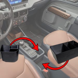 May include: Black plastic car console organizer with multiple compartments. The organizer is designed to fit in a car's center console. The image shows the organizer in a car interior with a steering wheel and seats.