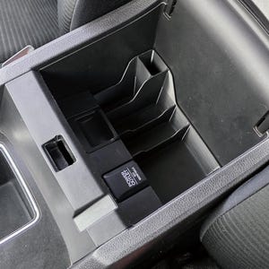 May include: Black car console with multiple storage compartments. Features a power outlet labeled "POWER OUTLET 12V MAX". The console is integrated into the vehicle's interior, with a textured fabric seat visible in the background.