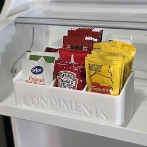 May include: A white plastic refrigerator shelf with a clear plastic lid, holding packets of condiments. The packets include ketchup, mustard, and crushed red pepper. The word "CONDIMENTS" is embossed on the shelf.