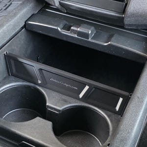 May include: Black car console with two cup holders and a small storage compartment. A rectangular tray with the text "AutoGadgets.com" is placed above the cup holders. The console is made of black plastic.