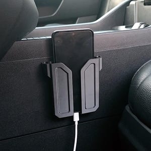 May include: Black car phone holder mounted on a car's interior panel. The phone is held in place by a custom-designed bracket. A white charging cable is plugged into the phone. The car's interior is black.