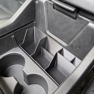 May include: Black car console organizer with cup holders and multiple storage compartments. The organizer is designed to fit inside a vehicle's center console, providing additional storage space for items like phones, keys, and other small objects.