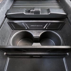 May include: Black car console with cup holders and storage compartments. The console includes a sliding tray and a compartment with the text "allweathercoating.com". The console is made of black plastic.