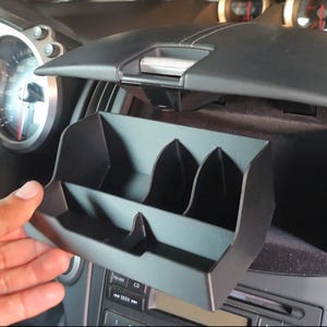 May include: A black car console organizer with three compartments is being held open. The organizer is designed to fit into a car's dashboard. The dashboard is black and has a speedometer and other controls visible.