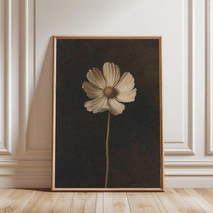Puede incluir: Impresión enmarcada de una flor blanca con centro amarillo sobre un fondo oscuro y texturizado. Los pétalos de la flor son detallados. El marco es de color marrón claro. La obra de arte está apoyada contra una pared blanca, creando una estética clásica.