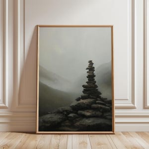 May include: Framed print depicting a stone cairn in a misty mountain scene. The image shows a carefully balanced stack of dark grey stones atop a larger rock formation. The background features a hazy, muted palette of greys and greens.