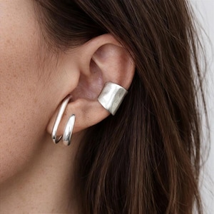 May include: A close-up of an ear displaying silver jewellery. The ear has a large, textured ear cuff and two curved hoop earrings. The jewellery has a polished, metallic finish, reflecting light. The person has brown hair.