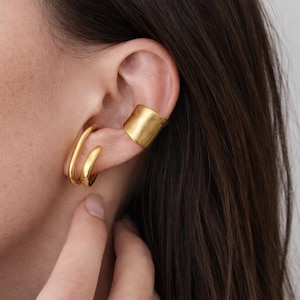 Könnte beinhalten: Goldfarbenes Ohrschmuck-Set. Das Set beinhaltet einen Doppelreifen-Ohrring und einen Ohr-Manschetten-Ohrring. Der Schmuck wird am Ohr einer Person mit dunkelbraunem Haar getragen. Die Ohrringe haben ein glänzendes, metallisches Finish.