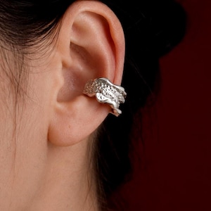 May include: A silver ear cuff with a textured, organic design. The cuff wraps around the ear and features small, sparkling embellishments. The jewelry is worn on the ear, adding a unique and stylish accent.