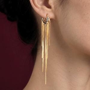 May include: Gold-toned earrings with a unique design. The earrings feature a curved top with a decorative element and long, dangling chains. The chains cascade down, creating a flowing, elegant look. The earrings are shown against a dark red background.