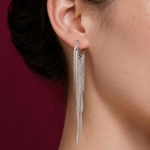 May include: Silver-toned hoop earrings with multiple dangling strands. The earrings are shown on a person's ear, with the strands cascading downwards. The design is sleek and modern, suitable for various occasions.