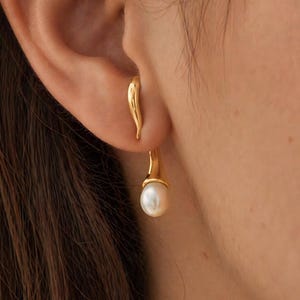 May include: A gold-toned ear cuff featuring a smooth, curved metal design that hugs the upper ear cartilage, connected to a dangling, teardrop-shaped white pearl pendant that hangs gracefully below the earlobe, creating a layered effect.