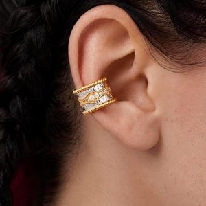 May include: A gold ear cuff featuring a wide band design with a textured beaded border. The center of the cuff is set with a row of baguette-cut clear stones arranged horizontally, accented by small round stones placed between the baguette sections.