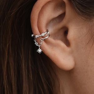 Puede incluir: Puño de oreja plateado con un diseño de doble banda. La banda superior tiene tres pequeñas piedras transparentes. Una cadena se conecta a un dije de estrella colgante. La joya se usa en la oreja.