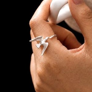 Op de afbeelding: Een zilverkleurige ring met een gestileerde vogel in vlucht met uitgestrekte vleugels. De ring is open aan de achterkant, waarbij de vleugels van de vogel het ontwerp van de band vormen. Het metaal heeft een matte afwerking en wordt gedragen.