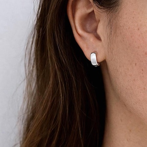 May include: A close-up of a silver hoop earring on a person's ear. The earring is a small, polished metal hoop with a flat front. The person has long, brown hair and fair skin.