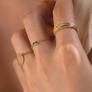 May include: Three gold rings worn on fingers. One ring features a textured herringbone pattern, another is a delicate chain ring with a small rectangular green stone, and the third is a thin chain ring with a small circular gold charm pendant.