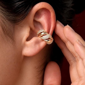 Pode incluir: Um ear cuff de tom dourado com um design torcido de várias camadas. A superfície da peça é adornada com inúmeras pedras pequenas, claras e cintilantes, cravadas em estilo pavé, criando uma aparência texturizada e reflexiva no metal.