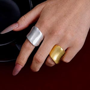 May include: A hand displays two chunky rings. One ring is silver, the other is gold. Both rings have a wide, irregular shape. The background is a dark red color, with a black bowl in the upper left corner.