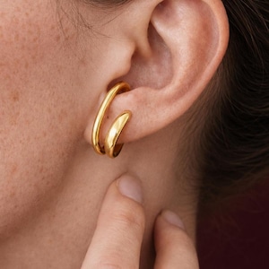 Op de afbeelding: Een close-up van een goudkleurige dubbele oorring. De oorring wordt gedragen op het oor van een persoon met donker haar. De oorring heeft een gladde, gepolijste afwerking en een gebogen ontwerp. De achtergrond is donkerrood.