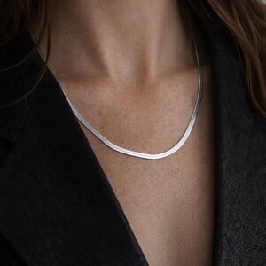 May include: A silver herringbone chain necklace. The necklace is flat and flexible, resting on a person's chest. The person is wearing a black jacket. The necklace is a simple, elegant piece of jewelry.