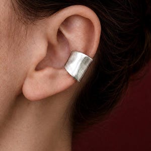 Pode incluir: Uma argola de orelha prateada é usada na orelha. A argola tem uma forma retangular com um design ligeiramente curvo. A joia é feita de um material metálico brilhante. O fundo é de cor vermelho escuro.