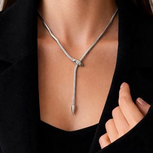 Può includere: Una collana argentata con un design a lariat unico. La collana presenta una catena a forma di serpente che culmina in un piccolo pendente rotondo con una pietra trasparente. Un pendente appeso e appuntito pende dal centro. La collana è indossata su un top nero.