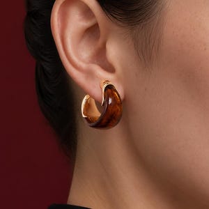 May include: A thick hoop earring featuring a polished gold-tone metal frame and a smooth, translucent amber-coloured resin insert with a marbled brown pattern. The earring has a curved, chunky silhouette that sits securely against the earlobe.