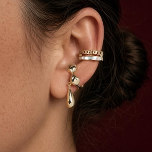 May include: Gold-toned jewelry set on an ear, including a cat-shaped earring with a teardrop pendant and two ear cuffs. One cuff has a chain design, and the other has a white band.
