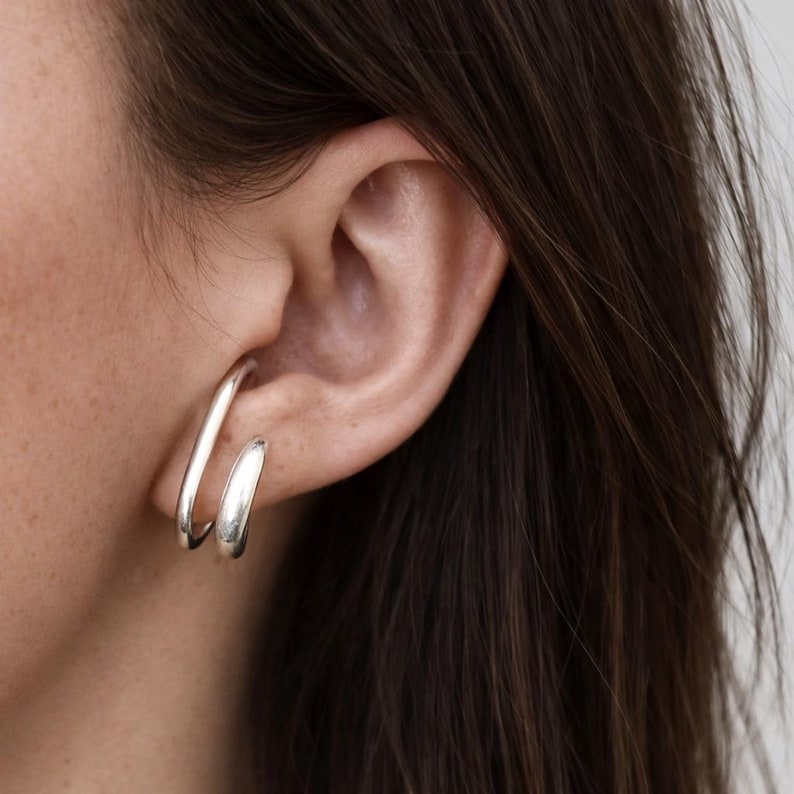 May include: A close-up of a silver earring set. The earrings are a modern design, with a curved bar that wraps around the ear and a small hoop. The earrings are made of a shiny, metallic material.