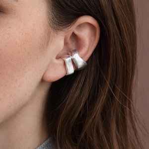 Puede incluir: Primer plano de un brazalete de oreja plateado. El brazalete presenta un diseño geométrico moderno con dos secciones distintas que se envuelven alrededor de la oreja. El metal tiene un acabado mate y la estética general es minimalista y contemporánea.