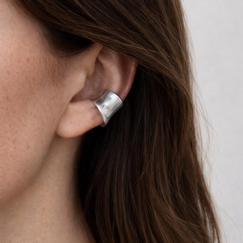 May include: A silver ear cuff with a curved, open design. The cuff wraps around the ear's cartilage, creating a modern and minimalist look. The metal has a slightly textured appearance.