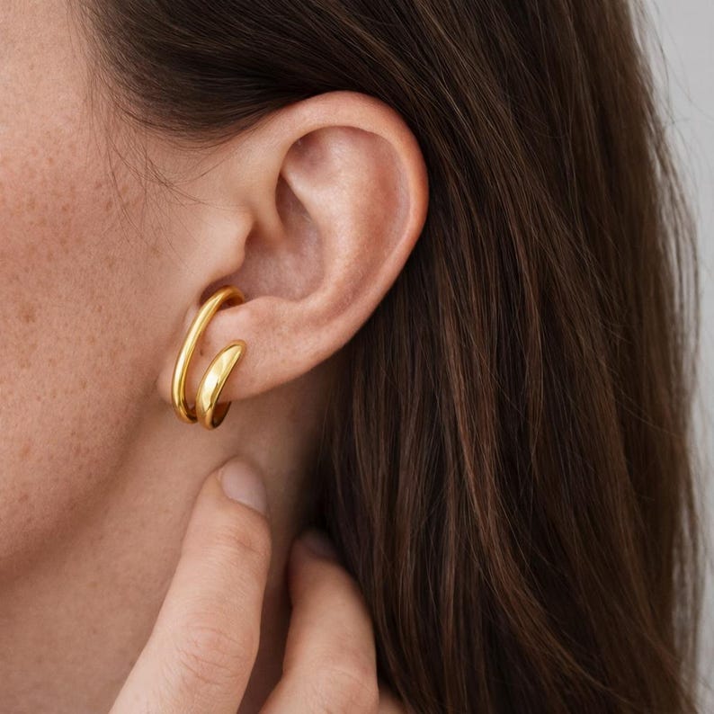 May include: A close-up of a gold-colored ear cuff. The cuff wraps around the ear with a curved design. The jewelry is shiny and reflects light. The person's ear and part of their hair are visible.