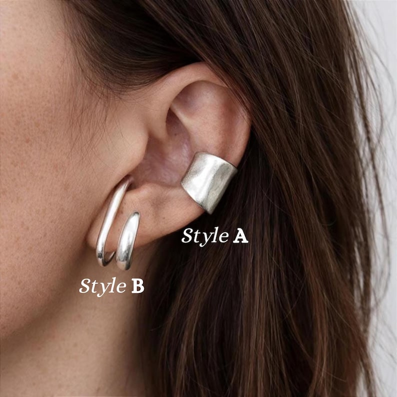 May include: Silver-toned ear jewelry, including a cuff labeled "Style A" and two hoop earrings labeled "Style B". The jewelry is worn on an ear, with the focus on the metallic accessories. The background is blurred, emphasizing the jewelry's design and texture.
