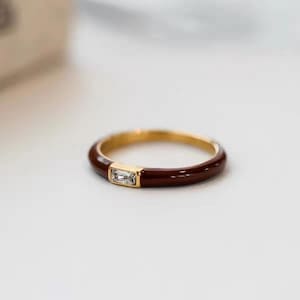May include: A gold ring featuring a dark red enamel band and a rectangular, clear gemstone. The ring is set against a white backdrop, accentuating its elegant design. The gemstone is set in a gold bezel.
