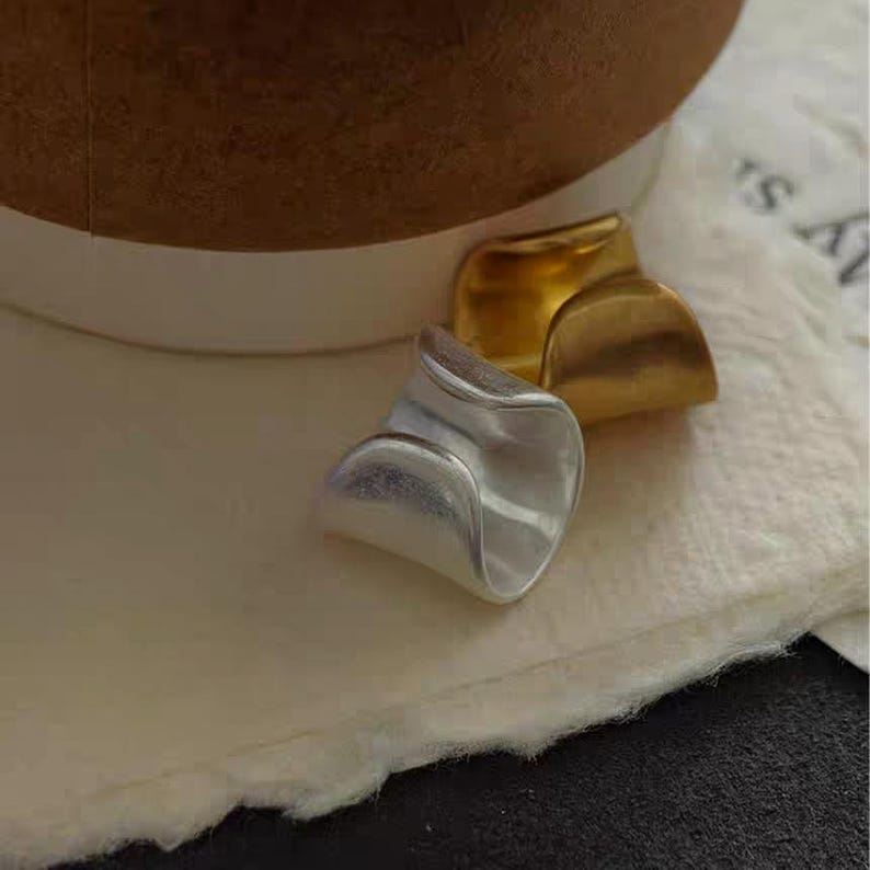 May include: Two rings, one silver and one gold, are displayed. The silver ring has a wavy, organic shape. The gold ring has a similar design, but is a different color. The rings are on a white surface.
