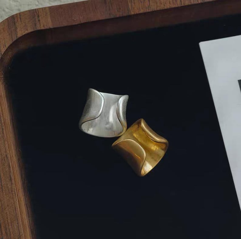 May include: Two abstract rings, one silver and one gold, rest on a black surface. The rings have a curved, geometric design. The silver ring is on the left, and the gold ring is on the right. The background is a dark wood tray.