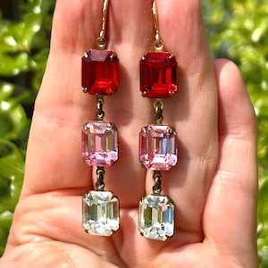 May include: A pair of drop earrings featuring three rectangular crystals in red, pink, and clear. The crystals are set in a gold-toned metal. The earrings are suspended from a gold-toned hook.