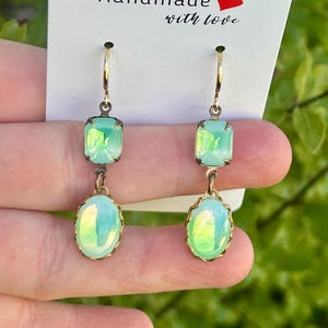 May include: A pair of dangle earrings with two green iridescent glass stones on each earring. The earrings are made with gold-toned metal.
