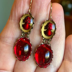 Könnte beinhalten: Ohrringe zum Hängen im Vintage-Stil. Jeder Ohrring hat ein rotes Cameo in einem goldfarbenen ovalen Rahmen, verbunden mit einem großen, ovalen, rubinroten Glasstein. Mit Hakenverschluss.