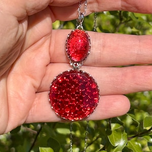 Op de afbeelding: Een zilveren ketting met twee rode glazen hangercharms. De grotere charm is rond en de kleinere is ovaal. Beide charms hebben een textuur en een glinsterende afwerking.