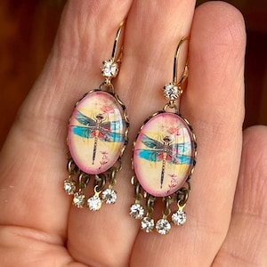 May include: Gold-tone dangle earrings featuring oval pendants with dragonfly illustrations. The earrings have a pink and yellow background with a dragonfly design. Each earring has a small crystal at the top and dangling crystals at the bottom.