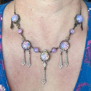 May include: A vintage-style necklace with iridescent, round, and oval-shaped stones in shades of purple and white. The necklace has a bronze-colored metal chain and decorative accents, including dangling pendants. The necklace is worn on a person.
