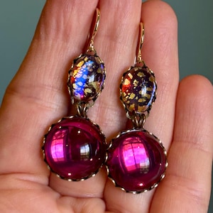 May include: A pair of dangle earrings with a gold-tone metal setting. Each earring features a small, oval-shaped top with blue, gold, and purple accents, and a large, round, magenta-colored bottom. The earrings are held by a hand.