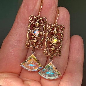 以下が含まれることがあります： アンティークゴールドトーンのイヤリングのペア。各イヤリングは、中央に虹色のクリスタルと、大きく三角形の虹色のクリスタルペンダントが付いた、華やかなフィリグリーデザインが特徴です。イヤリングは人の手で持たれています。