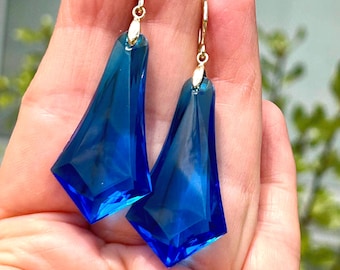 May include: A pair of blue, geometric earrings with gold-colored hooks. The earrings are a deep, transparent blue with a faceted design, catching the light. The earrings are held in a hand.