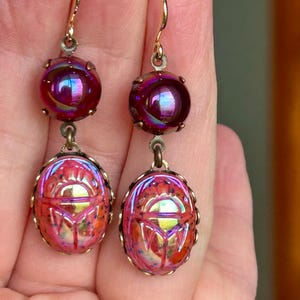 Può includere: Un paio di orecchini pendenti dall'estetica vintage. Ogni orecchino presenta una perla rotonda e iridescente viola nella parte superiore e un ciondolo a forma di scarabeo nella parte inferiore. I ciondoli sono un mix di rosa, arancione e giallo.