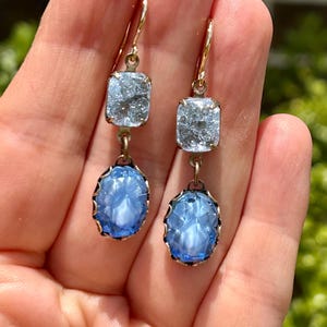 May include: A pair of dangle earrings with two blue glass stones on each earring. The top stone is a square shape with a crackled finish. The bottom stone is oval shaped with a carved design. The earrings are attached to gold-colored hooks.